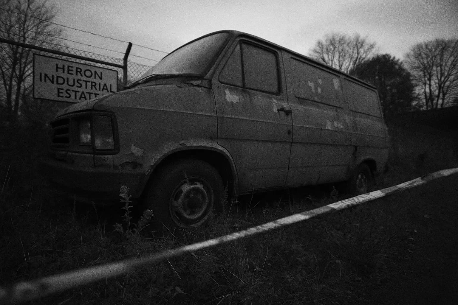 A gray van with peeling paint sits abandoned at Heron Industrial Estate, surrounded by overgrown weeds. Police tape flutters nearby, hinting at its connection to the unsolved Hammersmith Nude Murders.